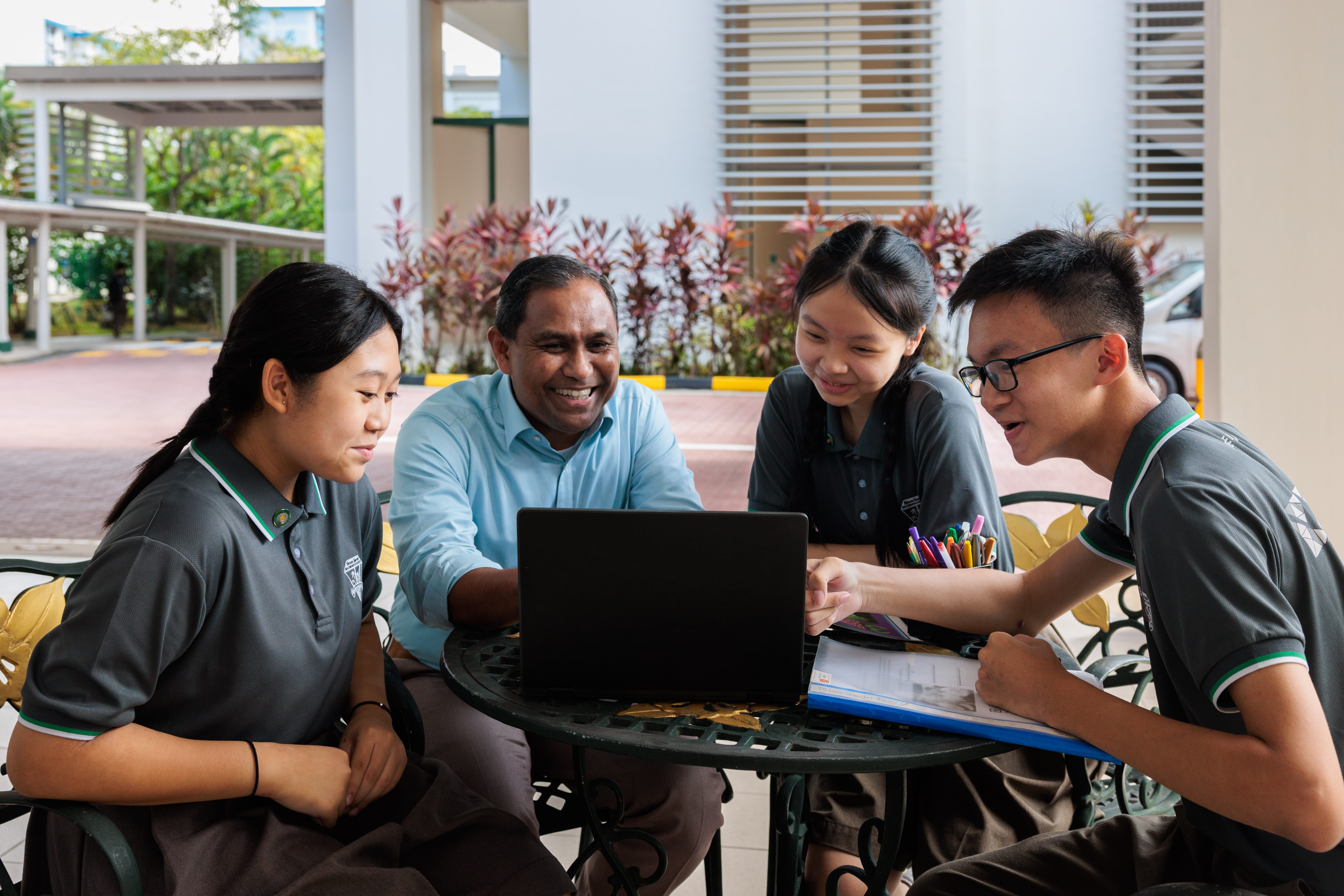 A smiling adult and three students in uniforms gather around a laptop at an outdoor table with notebooks and pens.